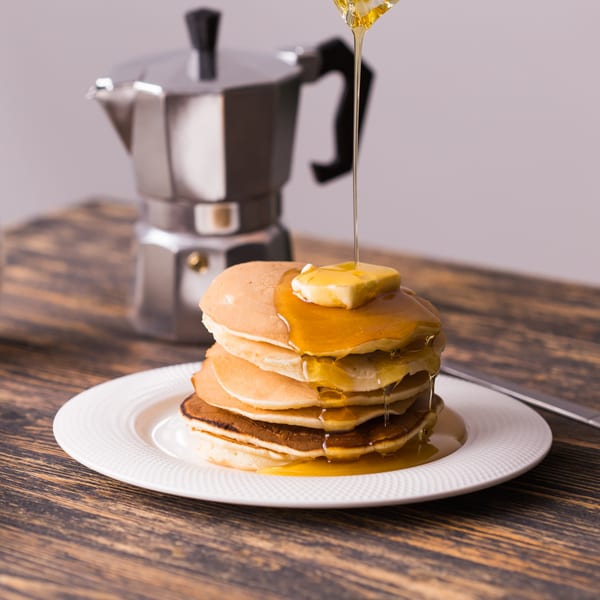 Syrup pouring over a stack of pancakes with butter on a white plate next to a moka pot.