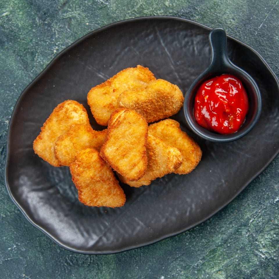 Golden-brown chicken nuggets on a black plate with a side of red ketchup.